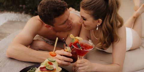 Ein lächelndes Paar in Badekleidung liegt auf einer Sonnenliege im Freien, starrt sich an und stößt mit einem Cocktailglas an. Ein Teller mit bunten Häppchen steht daneben. Die Stimmung ist entspannt und romantisch.