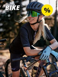 Eine Frau mit Helm, Sonnenbrille und Fahrradhandschuhen lehnt lächelnd im Freien auf einem Fahrrad, während das Sonnenlicht durch die Bäume fällt. Das Bild zeigt das Wort „BIKE“ und ein gelbes Prozentzeichen.
