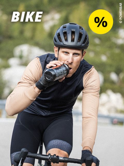 Ein Radfahrer mit Helm und Sportkleidung trinkt aus einer Flasche, während er im Freien Rad fährt. In der oberen linken Ecke erscheinen das Wort „BIKE“ und ein gelbes Prozentzeichen.