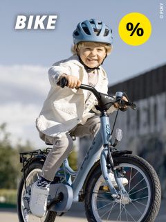 Ein lächelnder Junge mit Helm fährt an einem sonnigen Tag im Freien auf einem blauen Fahrrad. Das Wort „BIKE“ ist in fetten weißen Buchstaben geschrieben, und in der oberen rechten Ecke ist ein gelbes Prozentzeichen zu sehen.