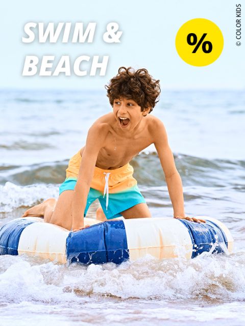 Ein lächelnder Junge in einer gelb-türkisen Badehose reitet auf einem blau-weißen aufblasbaren Ring im seichten Meerwasser. Der Text „SWIM & BEACH“ und ein gelbes Rabattsymbol sind zu sehen.