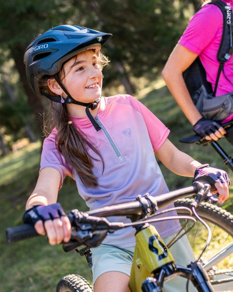 Ein junges Mädchen, das einen Helm und Handschuhe trägt, lächelt, während es den Lenker seines Fahrrads im Freien festhält; ein anderer Radfahrer steht neben ihr, teilweise sichtbar. Das Sonnenlicht fällt durch die Bäume im Hintergrund.