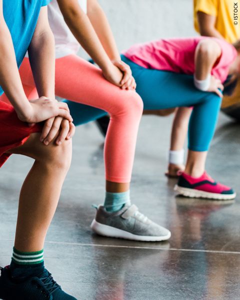 Eine Gruppe von Kindern in farbenfroher Sportkleidung dehnt sich, beugt sich mit den Händen auf den Knien nach vorne und bereitet sich auf eine körperliche Betätigung in der Halle vor.