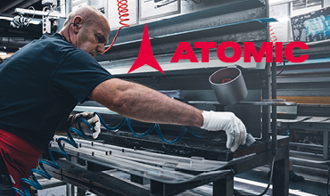 Ein Arbeiter in einer Werkstatt, bekleidet mit Handschuhen und roter Schürze, bedient Maschinen zur Skiherstellung. Das rote Atomic-Logo mit Dreieck ist sichtbar. Industrielles Umfeld mit verschiedenen Werkzeugen und Geräten.