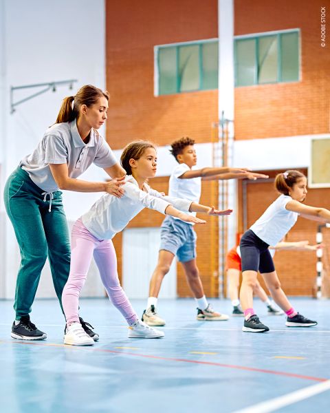Ein Trainer hilft einem jungen Mädchen bei einer Dehnungsübung in einer Turnhalle, während sich im Hintergrund andere Kinder in Sportkleidung dehnen.