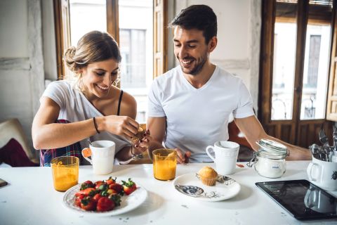 Ein lächelndes Paar sitzt an einem Küchentisch und frühstückt mit Kaffee, Erdbeeren, einem Muffin und Orangensaft. Sie wirken glücklich und entspannt in einer hellen, gemütlichen Umgebung mit großen Fenstern hinter ihnen.