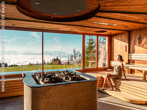 Eine Frau sitzt auf einer Holzbank in einer modernen Sauna mit großen Fenstern und genießt die Aussicht auf die Berge und das einfallende Sonnenlicht. Draußen sind Bäume und Wolken über einem grasbewachsenen Hang zu sehen.