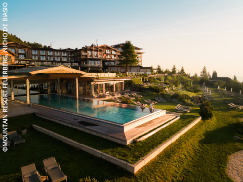 Ein luxuriöses Bergresort mit modernen Gebäuden, einem Infinity-Außenpool, Liegestühlen und angelegten Gärten auf einem Hügel, umgeben von Grün und einem klaren Himmel bei Sonnenuntergang.