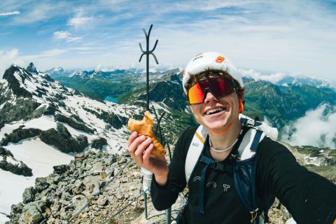 Ein lächelnder Bergsteiger mit Helm und Sonnenbrille macht ein Selfie auf einem verschneiten Berggipfel, in der Hand ein Gebäckstück. Majestätische Berge und Täler erstrecken sich im Hintergrund unter einem teilweise bewölkten Himmel.