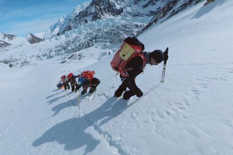 Eine Gruppe von Bergsteigern in Winterausrüstung wandert mit Trekkingstöcken einen steilen, verschneiten Berghang hinauf, mit schneebedeckten Gipfeln und Gletschern im Hintergrund unter einem strahlend blauen Himmel.