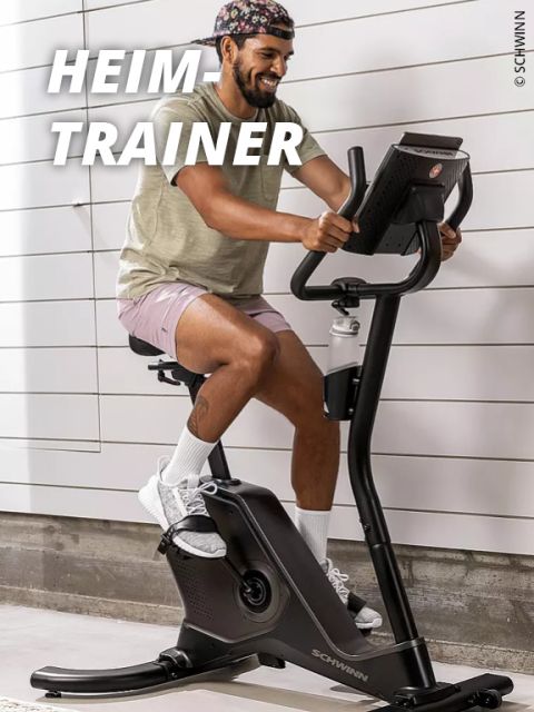 Ein lächelnder Mann mit Mütze, T-Shirt und Shorts fährt auf einem stationären Heimtrainer in einer Halle. Die Worte "HEIM TRAINER" erscheinen in großer weißer Schrift über ihm.