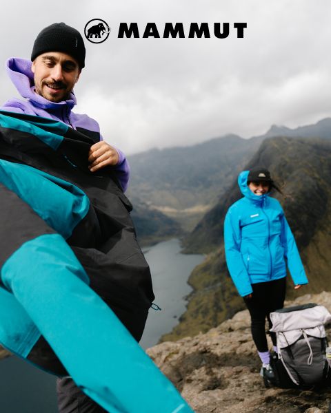 Zwei Wanderer in Outdoor-Ausrüstung durchqueren felsiges Gelände unter einem bewölkten Himmel, im Hintergrund ein großer Berg. Beide tragen Mammut-Kleidung, und das Markenlogo ist oben im Bild zu sehen.