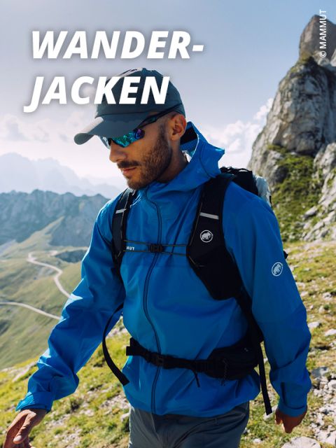 Ein Mann mit blauer Wanderjacke, Mütze und Sonnenbrille geht auf einem Bergpfad mit felsigen Gipfeln und einer kurvenreichen Straße im Hintergrund. Der Text "WANDER-JACKEN" erscheint oben links.