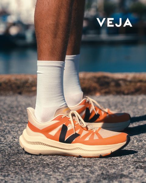 Eine Person trägt weiße Veja-Sneaker mit hellblauen Details und Gummisohle. Die Schuhe werden mit einer weißen Hose kombiniert. Der Hintergrund zeigt ein geometrisches Muster auf einer gefliesten Wand. Das Veja-Logo ist in der unteren rechten Ecke sichtbar.