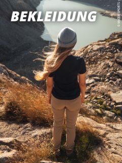 Eine Frau in Outdoor-Kleidung und mit einer Mütze steht auf einem felsigen Berg und blickt auf einen See. Der Text oben lautet "BEKLEIDUNG" in fetten, weißen Buchstaben.