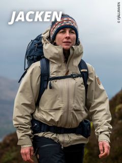 Eine Person, die eine beige Outdoor-Jacke, eine bunte Strickmütze und einen Rucksack trägt, wandert in einer bergigen Landschaft. Das Wort "JACKEN" erscheint in fetter weißer Schrift oben links.