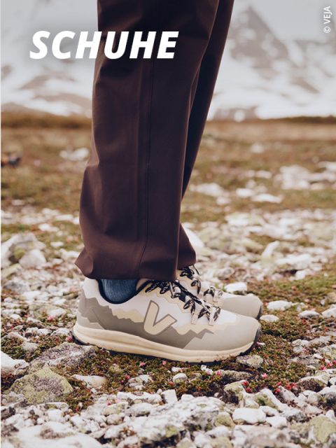 Eine Person steht im Freien auf felsigem Grund und trägt eine braune Hose und beigefarbene Wanderschuhe mit einem Bergmuster. Im unscharfen Hintergrund sind schneebedeckte Berge zu sehen. Das Wort "SCHUHE" erscheint in fetter weißer Schrift.