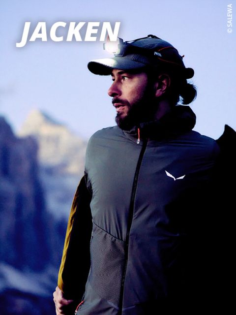 Ein Mann mit Stirnlampe und Outdoor-Jacke blickt in der Abenddämmerung in einer bergigen Gegend zur Seite. Das Wort "JACKEN" ist in fetter weißer Schrift oben links zu sehen.