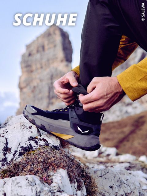 Eine Person in schwarzen Wanderschuhen zieht die Schnürsenkel fest, während sie auf felsigem Gelände steht, mit einem Berggipfel und dem Wort "SCHUHE" in fetten weißen Buchstaben im Hintergrund.