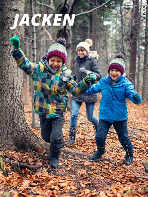 Zwei Kinder in bunten Winterjacken und Mützen laufen durch einen Wald mit gefallenen Blättern, halten sich an den Händen und lächeln, während ein Erwachsener in einer dunklen Jacke hinterherläuft. Das Wort "JACKEN" erscheint in fetter weißer Schrift.