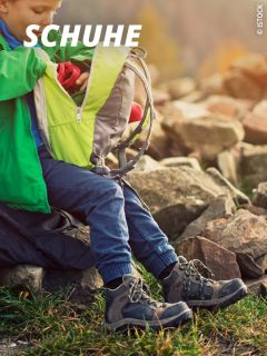 Ein Kind in Wanderschuhen und blauer Hose sitzt auf einem Felsen im Freien und öffnet einen grün-grauen Rucksack. Oben links ist in weißer Schrift das Wort "SCHUHE" zu lesen.