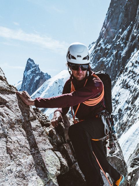 Ein Bergsteiger mit Helm, Sonnenbrille und Kletterausrüstung erklimmt einen felsigen Berghang mit schneebedeckten Gipfeln im Hintergrund unter einem klaren Himmel.