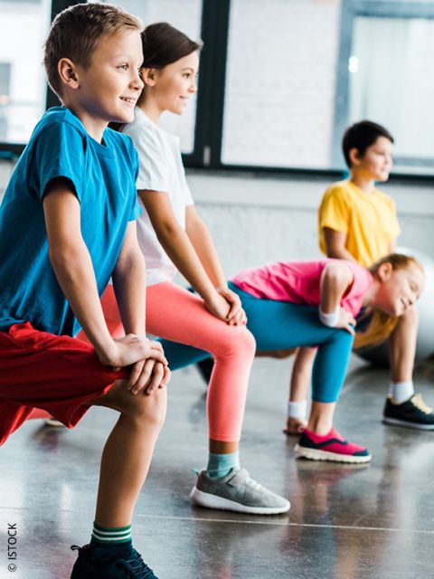 Vier Kinder in farbenfroher Sportkleidung stehen in einem Raum und machen eine Dehnungsübung mit den Händen auf den Knien, lächeln und schauen nach vorne. Der Raum hat große Fenster und einen glänzenden Boden.