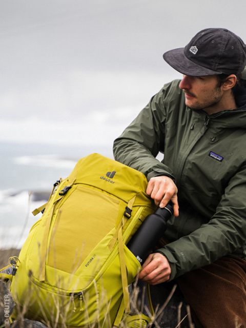 Eine Person mit dunkler Mütze und grüner Jacke sitzt im Freien, hält einen gelben Deuter-Rucksack und steckt eine schwarze Wasserflasche in dessen Seitentasche. Der Hintergrund ist wolkenverhangen und lässt einen Hauch von Wasser und Gras erkennen.