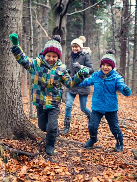 Zwei Kinder in bunten Jacken und Mützen rennen und spielen fröhlich in einem Wald mit Herbstlaub, während ein Erwachsener lächelnd hinterherläuft. Alle sind warm gekleidet für das kalte Wetter.
