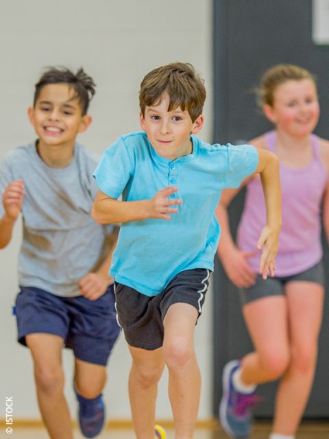 Drei Kinder in Sportkleidung laufen im Haus, lächeln und scheinen voller Energie zu sein. Der Fokus liegt auf dem Jungen in der Mitte, der ein hellblaues Hemd trägt, während zwei andere Kinder im Hintergrund leicht verschwommen sind.