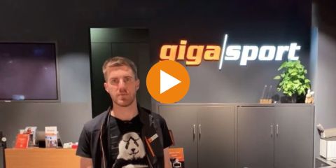 Eine Person steht in einem schwach beleuchteten Raum vor einer Wand mit dem Logo „gigasport“. Oben ist der Text „E-Lastenrad Tipp“ zu sehen. In der Bildmitte befindet sich ein großer orangefarbener Play-Button.