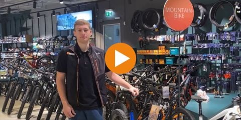 Eine Person steht in einem Fahrradladen neben einer Reihe Mountainbikes. Im Hintergrund ist verschiedenes Fahrradzubehör zu sehen. Oben erscheint der Text „E-MOUNTAINBIKE TIPP“, und mittig im Bild befindet sich ein Play-Button.