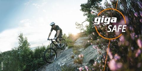 Ein Radfahrer mit Helm fährt einen steinigen Weg entlang, der von Bäumen und Blumen umgeben ist. Der Himmel ist teilweise bewölkt. Der Text lautet „Sicherheit beim Radfahren“ und „giga SAFE“ mit einem kreisförmigen Design.
