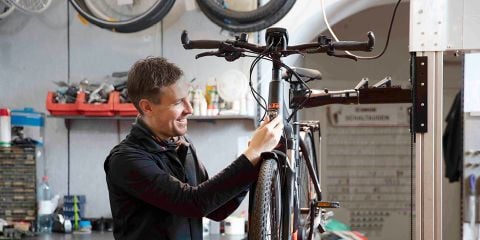 Eine Person wartet ein Fahrrad in einer Werkstatt voller Werkzeuge und Fahrradteile. Der Text lautet „Fahrrad- und E-Bike-Service“. Im Hintergrund sind an den Wänden verschiedene Fahrradräder zu sehen. Die Person scheint die Vorderseite des Fahrrads einzustellen.