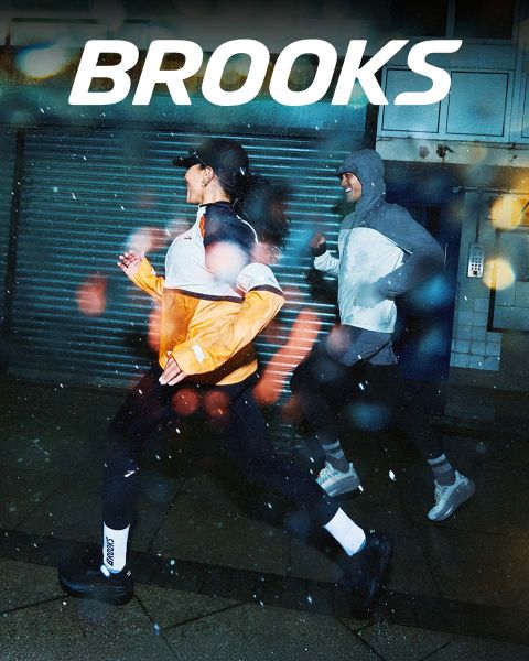 Eine Gruppe von fünf Personen rennt energisch über einen Zebrastreifen. Sie tragen farbenfrohe Sportkleidung, darunter Shorts und Tanktops. Die Szene ist lebhaft, über ihnen prangt deutlich das Wort „BROOKS“.