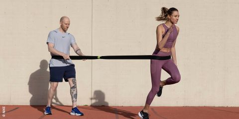 Eine Frau in Sportkleidung läuft mit einem Widerstandsband vorwärts, das von einem tätowierten Mann in Sportkleidung gehalten wird. Der Text lautet „Workout mit Matthias Snobe“ auf beigem Hintergrund.