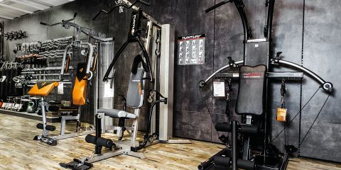 Ein Fitnessgeräte-Showroom mit verschiedenen Trainingsgeräten. Zu den Geräten gehören Hantelbänke, Kabelzugmaschinen und eine Kraftstation. In der oberen linken Ecke ist der Schriftzug „MONTAGE-SERVICES“ zu sehen. Der Boden ist aus Holz, die Wände dunkelgrau.