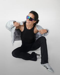 Eine Frau sitzt auf dem Boden und zieht sich lächelnd ein graues Sweatshirt über den Kopf. Sie trägt ein schwarzes ärmelloses Oberteil, eine graue Jogginghose, weiße Socken und graue Turnschuhe vor einem schlichten hellgrauen Hintergrund.