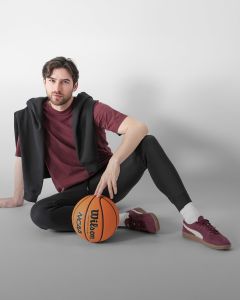 Ein Mann mit braunem Haar und Bart sitzt auf dem Boden vor einem weißen Block. Er trägt eine hellgraue Jacke, ein schwarzes Hemd, dunkle Jogginghosen, weiße Socken und graue Turnschuhe und hält eine Wasserflasche in der Hand.