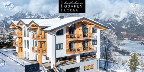 In einer verschneiten Berglandschaft steht eine moderne, mehrstöckige Hütte mit Holzbalkonen und weißer Fassade. Auf einem Schild steht "Premium Hapfelmeier Gumpen Lodge". Im Hintergrund sind Bäume und ferne Berge zu sehen.