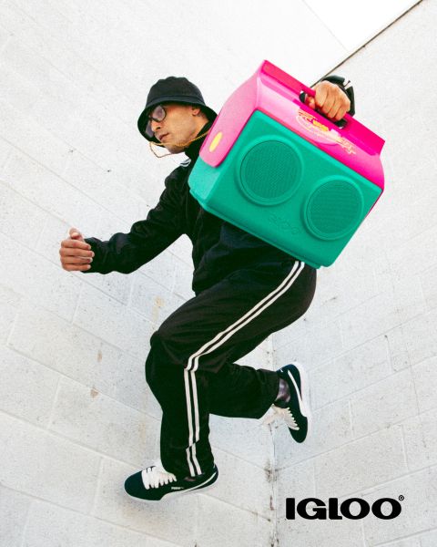 Eine lächelnde Frau mit lockigem Haar trägt eine pfirsichfarbene Igloo-Kühlbox an einem Seilgurt über der Schulter. Sie trägt eine weiße Kurzarmbluse und hellblaue Jeans und steht vor einem hellpfirsichfarbenen Hintergrund.