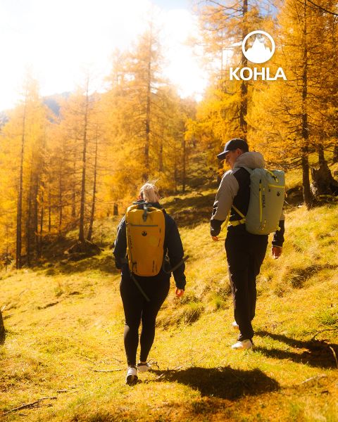 Zwei Wanderer mit Rucksäcken gehen durch einen sonnenbeschienenen Wald mit goldenen Herbstbäumen. Das Sonnenlicht fällt durch die Äste und wirft ein warmes Licht. Das Kohla-Logo ist in der oberen rechten Ecke zu sehen.