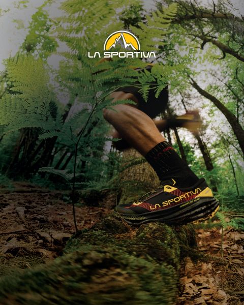 Vier Personen in Outdoor-Ausrüstung und mit Bouldermatten auf dem Rücken wandern einen grasbewachsenen Bergpfad hinauf. Über ihnen ist das Logo von La Sportiva am Himmel zu sehen. Im Hintergrund sind schroffe Berggipfel zu sehen.