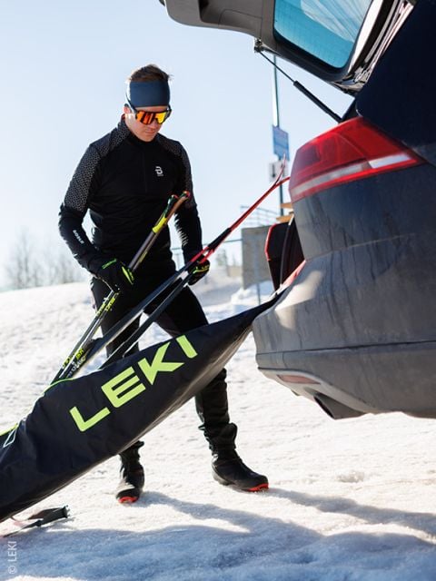 Eine Person in Wintersportkleidung und mit Sonnenbrille lädt an einem verschneiten Tag Langlaufskier aus einer Tasche mit der Aufschrift "LEKI" in den offenen Kofferraum eines Autos ein.