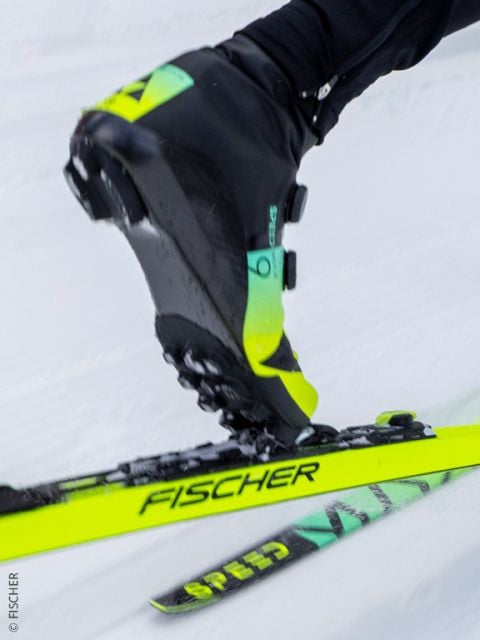 Nahaufnahme des Skischuhs einer Person, der auf einen leuchtend gelben Fischer-Langlaufski geschnallt ist, der über den Schnee gleitet. Sowohl der Schuh als auch der Ski tragen deutlich das Fischer-Logo.