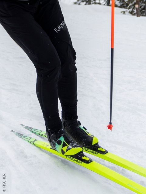 Nahaufnahme einer Person beim Skilanglauf auf Schnee, die ihre Unterschenkel und Füße zeigt. Sie trägt eine schwarze Hose und neongelbe Fischer-Ski, rechts ist ein Skistock zu sehen.