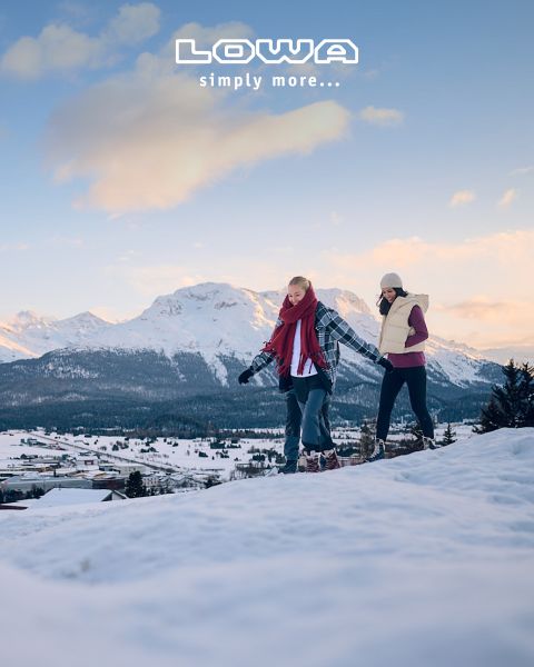 Drei Personen wandern mit Trekkingstöcken und Outdoor-Ausrüstung auf einem malerischen Bergpfad. Im Hintergrund sind Bäume und Berge zu sehen. Der Himmel ist bewölkt. Oben steht der Schriftzug „LOWA simply more…“.
