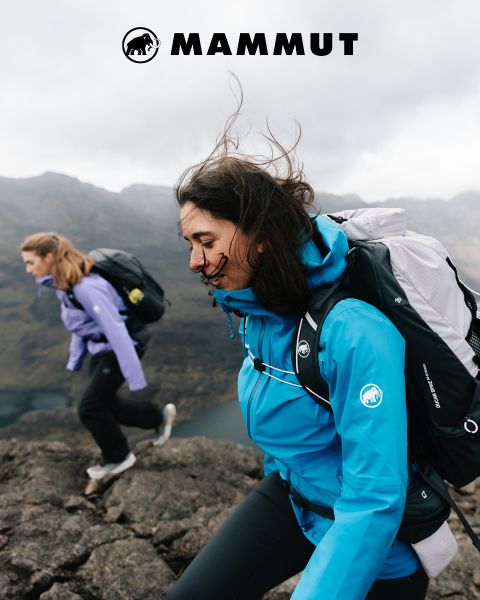 Zwei Frauen in Outdoor-Ausrüstung und Rucksäcken wandern in felsigem Gelände mit Bergen und bewölktem Himmel im Hintergrund. Das Mammut-Logo erscheint am oberen Rand des Bildes.