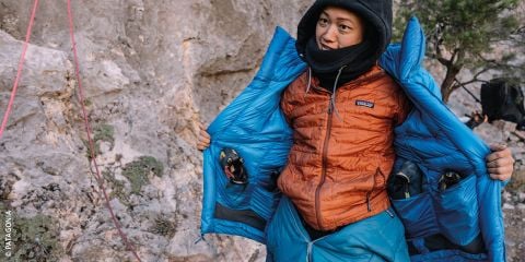 Eine Person im Freien trägt eine orangefarbene Daunenjacke, die geöffnet ist und eine blaue Jacke darunter zum Vorschein bringt. Sie richtet ihre Kleidung, umgeben von felsigem Gelände und Kletterausrüstung. In weißem Text steht „DAUNENJACKEN PFLEGEN“ mit einem Logo auf der rechten Seite.