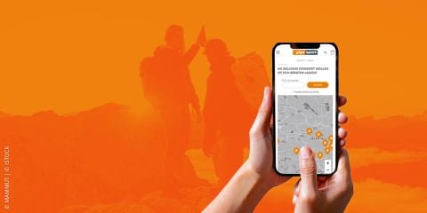 Eine Person hält ein Smartphone, auf dem eine Karte mit Standortmarkierungen angezeigt wird. Im Hintergrund sind Silhouetten von Wanderern auf einem Berggipfel in Orangetönen zu sehen. Der Text auf dem Bild lautet „Gigasport Sportberatung“.
